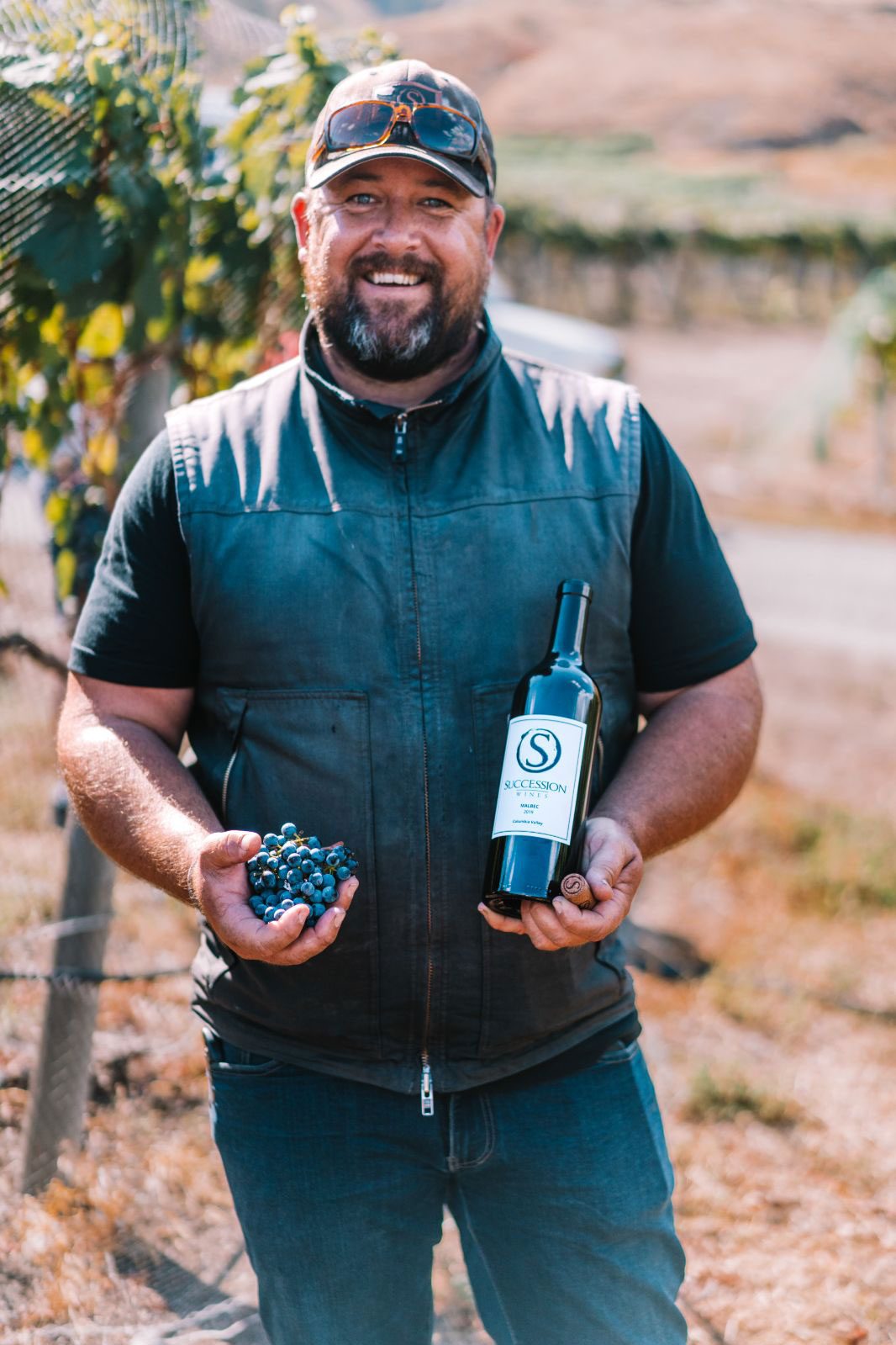 Brock Lindsay holding grapes and a bottle of wine in the vineyard